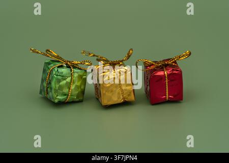 Three tiny gifts are arranged side by side on a green surface. They are wrapped in shiny paper with ribbons. The colors of the gifts are green, gold, Stock Photo