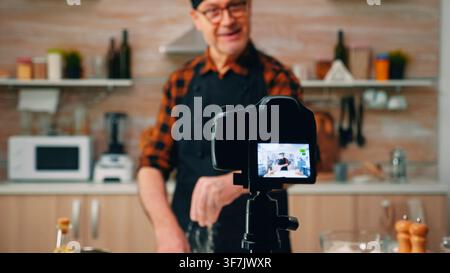 In a domestic kitchen, the male baker records a baking demonstration for online viewers as the recording is prepared for a live online audience. Stock Photo