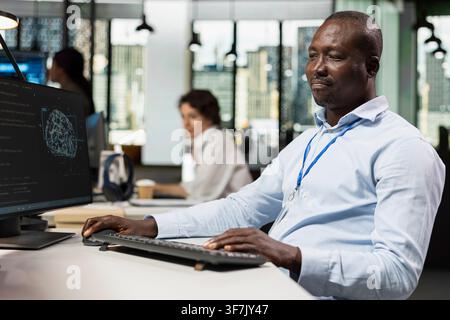 Programmer in small business workspace using computer to oversee artificial intelligence neural networks. African american man in tech startup office using AI machine learning systems on PC Stock Photo