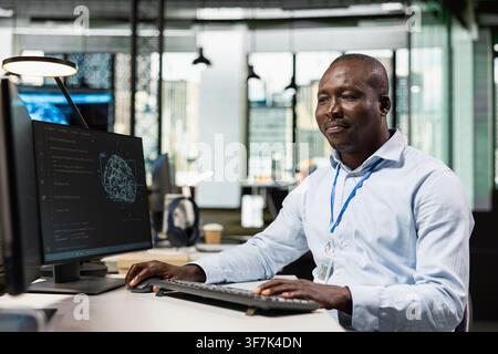 Smiling programmer in AI tech company workplace using PC to conduct code reviews. Happy IT professional using computer program at office desk to design and implement machine learning algorithms Stock Photo