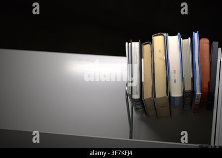 A row of books stands neatly arranged on a white shelf against a dark background. Stock Photo