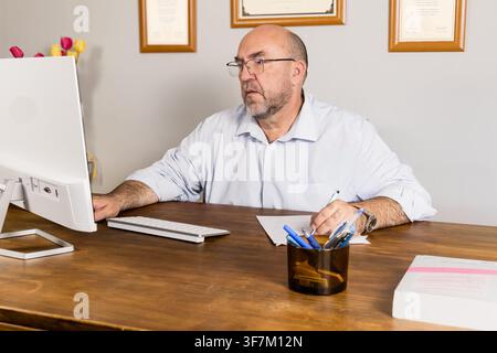 Middle-aged businessman in glasses working at a desktop computer with papers and pen on a wooden desk. Ideal for office work, business management and Stock Photo