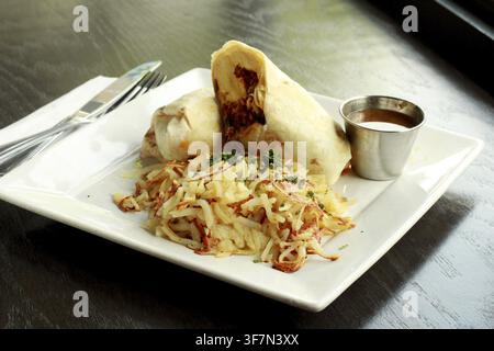 A breakfast burrito served on a square white plate with hash browns and a side of sauce Stock Photo