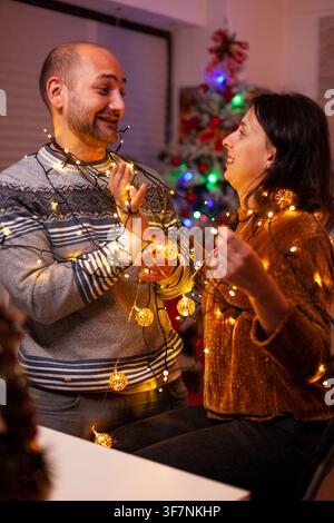 Glowing tree lights wrap around partners as they prepare seasonal kitchen decorations and enjoy the winter holiday. Stock Photo