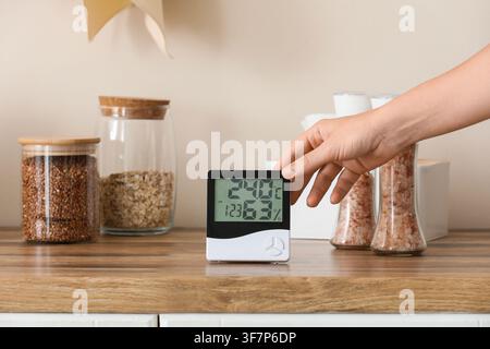 Womans hand adjusting a digital thermometer and hygrometer displaying temperature and humidity in a kitchen setting, the device displays indoor temper Stock Photo