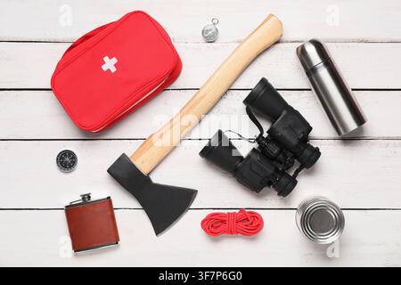 Overhead view of essential camping and survival gear including a first aid kit axe and binoculars on white wooden surface Stock Photo