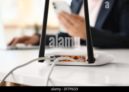 White wireless router with ethernet cables connected on a desk, person using laptop and smartphone in background, internet connection Stock Photo