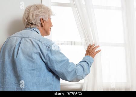 Senior woman with short white hair looking out a window, holding back a sheer white curtain, from behind Stock Photo