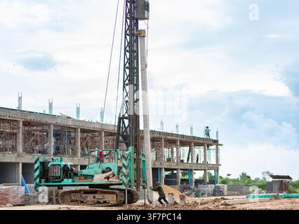 Construction workers operating a crawler pile driver machine to set up concrete foundation pillars near a building under construction. Stock Photo