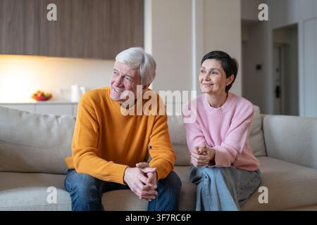 Happy senior married couple husband and wife sitting couch at home looking away at window. Stock Photo
