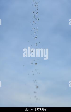 Droplets fall slowly against a clear blue sky. Each drop reflects the soft daylight above. Faint clouds drift in the background. Perfect for calm, nat Stock Photo