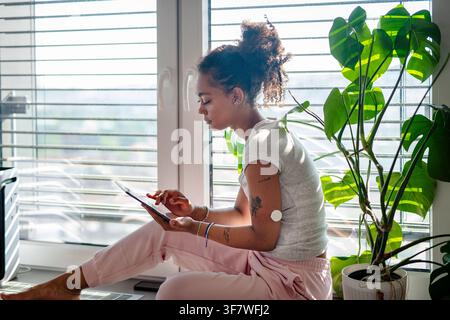 Female diabetes patient with CGM sensor using tablet in bright apartment Stock Photo
