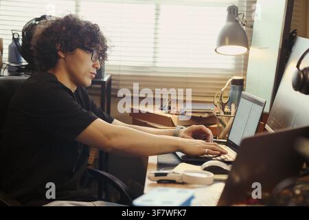 Asian young man typing on laptop at home desk, coding and working on software development. Image suitable for remote work, IT education, and freelance programming campaigns Stock Photo