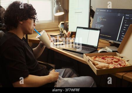 Young Asian man drinking beverage while coding on laptop and computer screens at home desk with pizza box, representing remote software developer lifestyle, medium shot Stock Photo