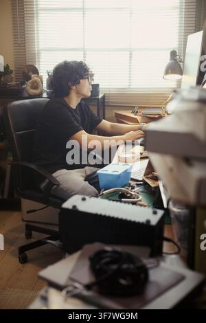 Vertical image of Young Asian man sitting at desk and typing on computer keyboard. Scene represents remote work or freelance programming, ideal for tech career marketing Stock Photo