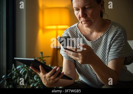 Mature woman using a digital tablet and smartphone to synchronize data between devices at home. Cross-device connectivity, cloud technology and digita Stock Photo