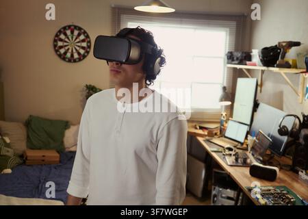 Young Asian man wearing virtual reality headset and headphones in home office, experiencing immersive digital simulation, ideal for tech marketing and gaming industry ads Stock Photo