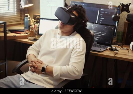 Young Asian man wearing virtual reality headset while sitting at desk with multiple computer screens. Image represents digital life and is suitable for tech marketing Stock Photo