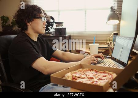 Asian young man working on laptop at home desk with pizza box in foreground, representing remote work lifestyle. Image suitable for food delivery and freelance campaign Stock Photo