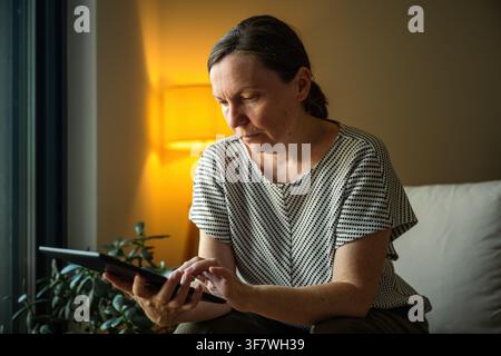 Woman using a tablet computer in a comfortable home setting during the evening. Digital lifestyle, remote work and online communication concept. Selec Stock Photo
