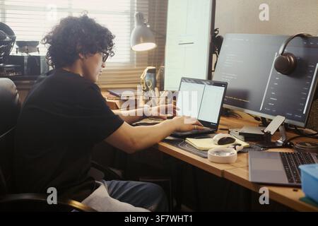 Young Asian man coding on laptop at home office desk with multiple monitors. Image represents remote software development, IT freelancing, and digital technology careers Stock Photo