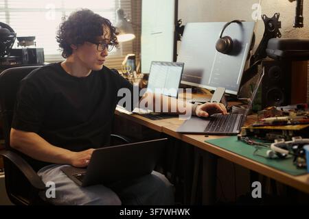 Asian young man working on multiple laptops at home office desk, coding and testing hardware. Image suitable for remote work, software development, and IT education campaigns Stock Photo
