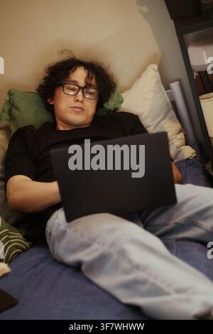 Vertical image of Young Asian man lying on bed and typing on laptop, working remotely from home. Concept of digital nomad lifestyle, suitable for remote work marketing campaigns Stock Photo