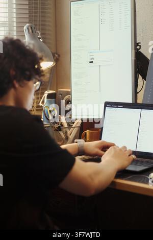 Young Asian man coding on laptop with vertical monitor showing code. Image ideal for software development, remote IT work, and programming education campaigns, vertical Stock Photo