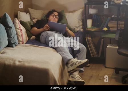 Asian young man lying on bed and using digital tablet in bedroom, studying or relaxing. Image suitable for technology, remote learning, and digital lifestyle marketing campaigns Stock Photo