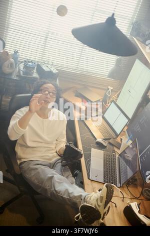 Vertical image of Asian young programmer throwing baseball at home office desk, taking break from coding. Concept of work life balance, suitable for remote work marketing Stock Photo