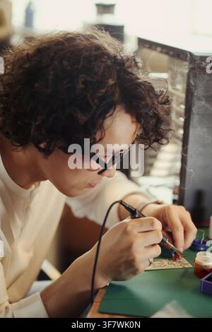 Vertical image of Asian young man soldering circuit board in workshop, focusing on hardware engineering. Image represents STEM education and technology development marketing Stock Photo