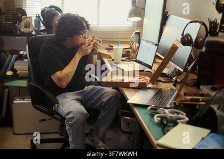 Young Asian man eating pizza at desk with multiple computers, taking break from coding. Image represents remote work lifestyle, suitable for tech industry recruiting campaigns Stock Photo