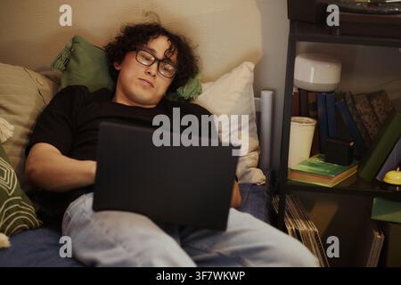 Asian young man lying in bed and using laptop computer, studying or working remotely. Image suitable for remote work, online education, and digital lifestyle marketing campaigns Stock Photo