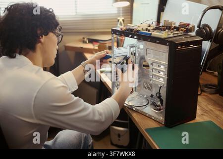 Young Asian man repairing desktop computer at home workshop, installing cooling fan. Image suitable for IT support services, hardware maintenance, and technology education Stock Photo