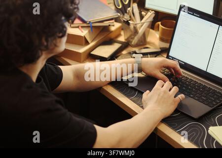 young man typing on laptop keyboard at wooden desk, writing software code. Over shoulder view representing remote work, programming education, and software development services Stock Photo