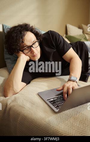 Vertical image of Young Asian man lying on bed and typing on laptop. Image represents remote work, online education, or digital lifestyle for e-commerce marketing Stock Photo