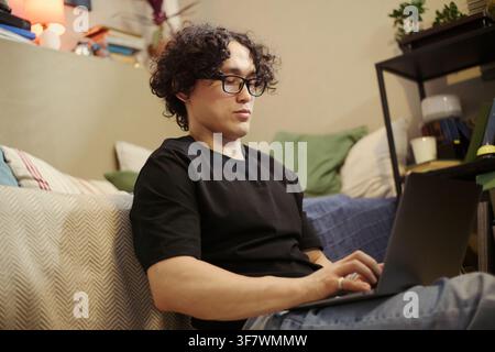 Asian young man wearing glasses typing on laptop while sitting on floor in living room, working remotely. Image suitable for remote work and online education marketing campaigns Stock Photo
