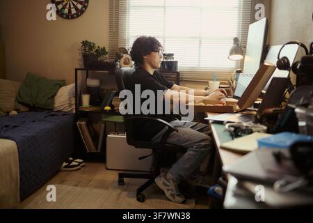 Young Asian man working on computer in bedroom, typing on keyboard and eating pizza. Image represents remote work, freelance lifestyle, and student housing marketing Stock Photo