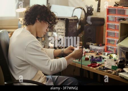 Young Asian man soldering circuit board at workshop desk, repairing computer hardware. Image represents engineering education, tech repair services, and DIY electronics hobby Stock Photo