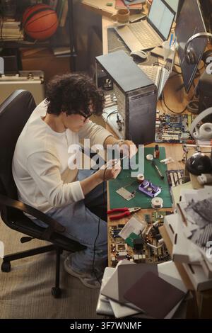 Young Asian man soldering microchip at messy desk, repairing computer hardware. High angle view representing technology education and IT support services, vertical Stock Photo