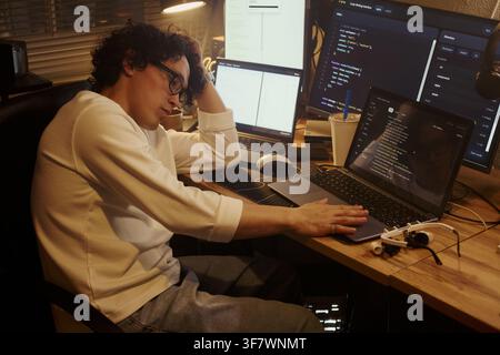 Tired Young Asian man sitting at desk with multiple monitors, coding on laptop and looking exhausted. Image represents developer burnout, suitable for IT career articles Stock Photo