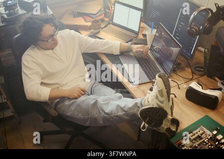 Young Asian man coding on laptop with feet on desk, working in home office. High angle view of software developer programming, suitable for remote work and IT career concepts Stock Photo