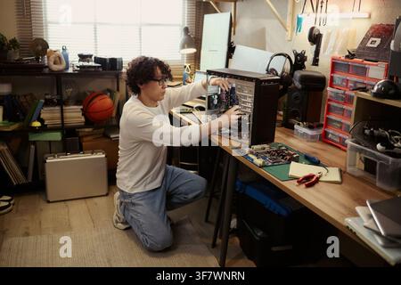 Young Asian man repairing desktop computer hardware in home workshop. Image represents IT support, technology maintenance, and DIY electronics repair for tech blog use Stock Photo