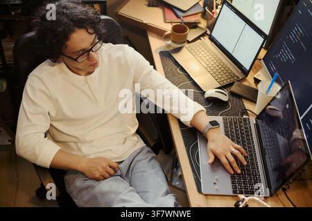 Young Asian man writing code on laptop at desk with multiple screens, representing software development and remote work. High angle shot Stock Photo