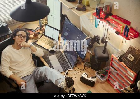Asian young man sitting at messy desk with multiple screens, coding and thinking. Image represents remote work, software development, and IT education, high angle shot Stock Photo