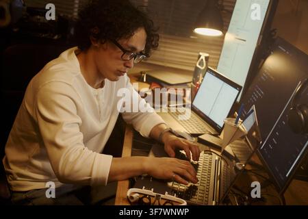 Asian young man typing on laptop at desk with multiple screens, coding software at night. Image represents remote work and IT development, suitable for tech recruitment ads Stock Photo
