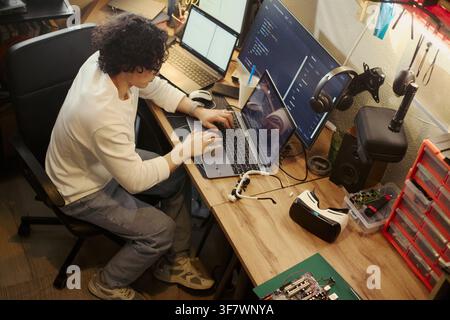 Young Asian man coding on laptop at messy desk with multiple screens, developing software. Image represents remote work and tech education, suitable for IT recruitment ads, high angle shot Stock Photo
