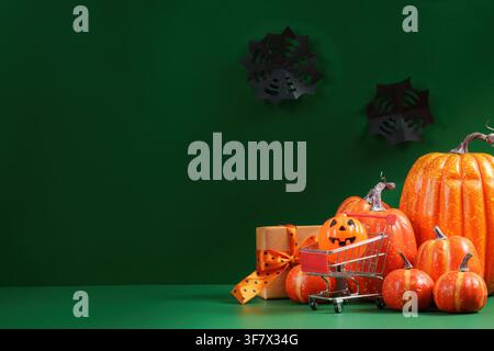 Halloween shopping concept with pumpkins, gift box, and mini shopping cart on green background Stock Photo