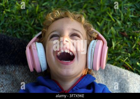 Smiling toddler child lying on green grass, wearing pink headphones, experiencing pure happiness and fun Stock Photo