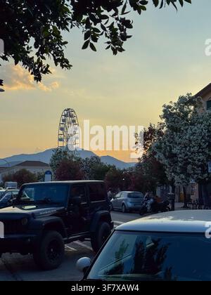 Tortoli, Italy, 20.06.25 Ferris wheel silhouetted at sunset behind trees and parked cars in a Sardinian town, with mountains in the background. Stock Photo